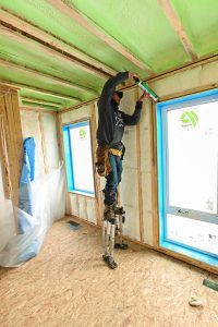 construction worker on stilts applies sealant to the ceiling of a new Avalon Homes property under construction, with green spray foam insulation visible on the ceiling and walls.