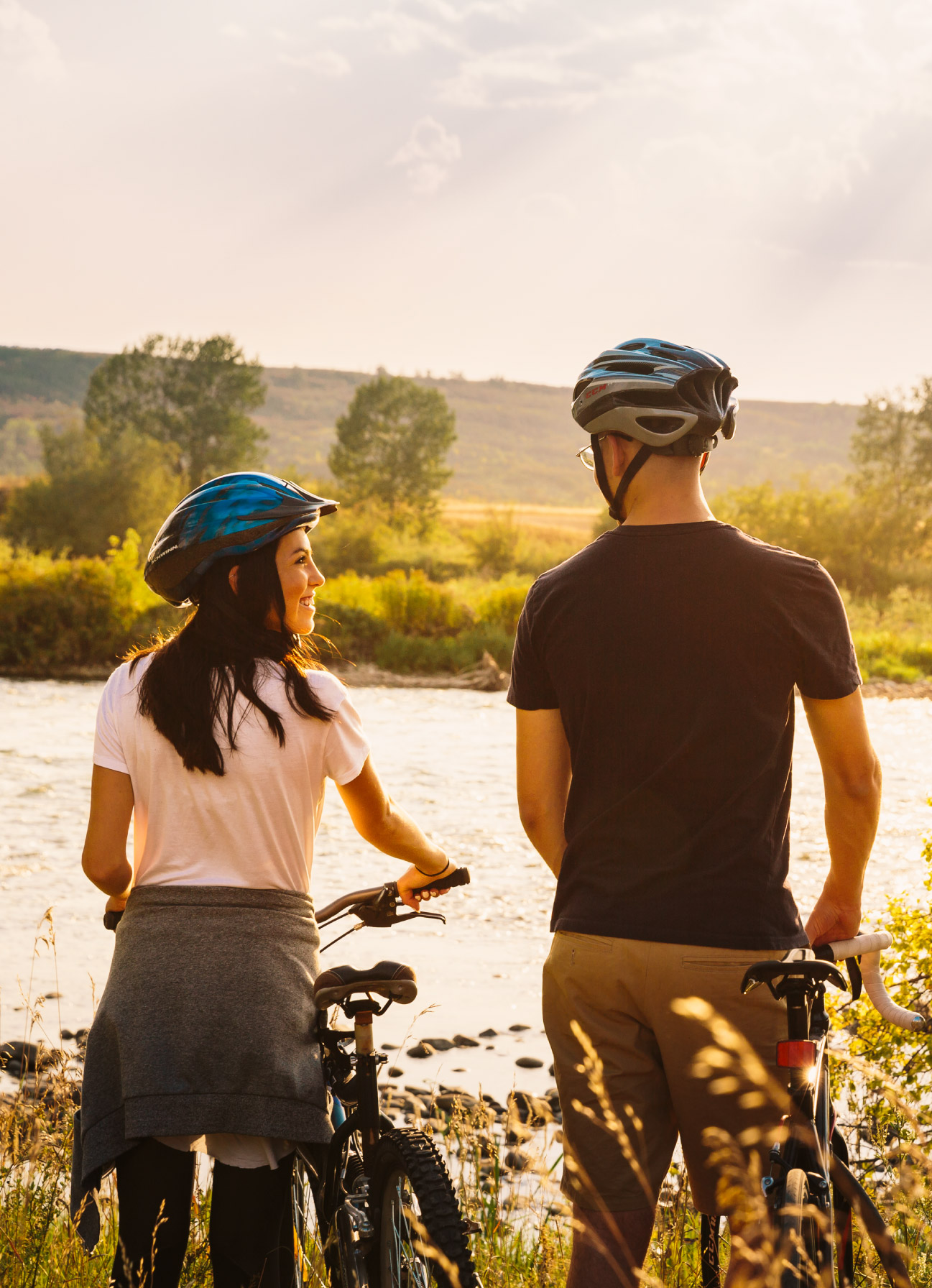 A couple standing next to their bicycles looking at each other facing a river at sunset