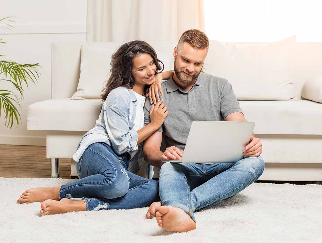couple sitting on floor in front of a white couch looking at laptop