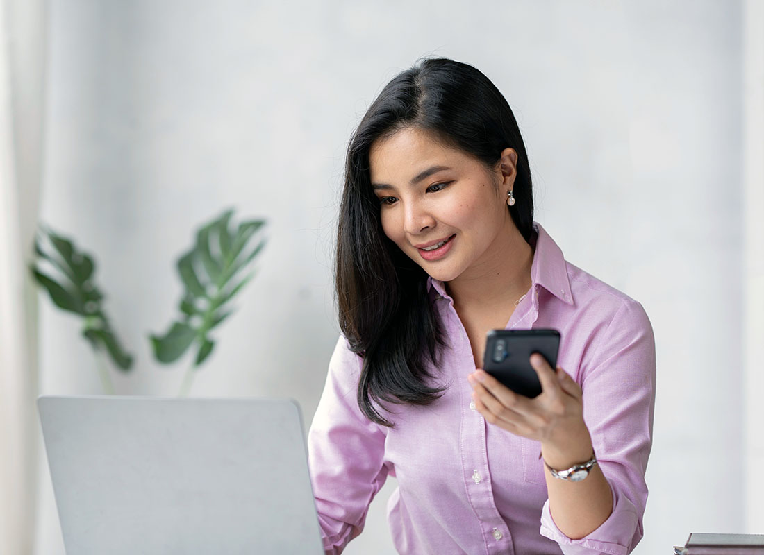 woman looking at laptop and holding a black cell phone in her left hand in a white room