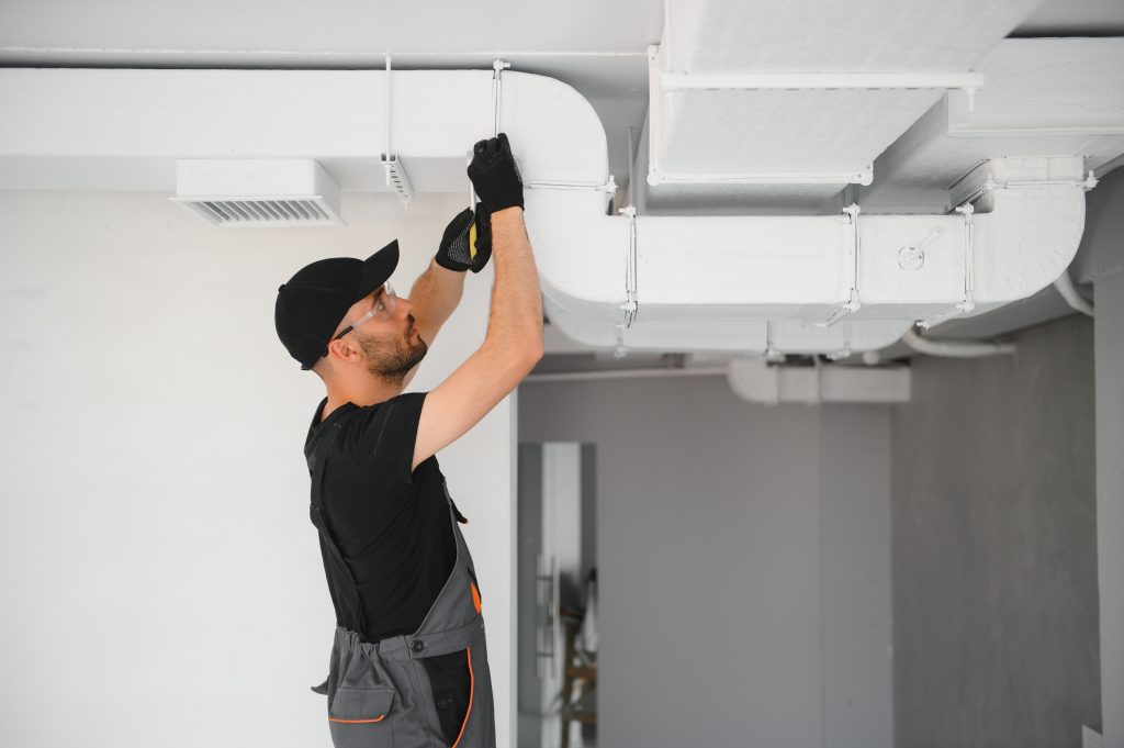 HVAC technician in black uniform and cap installing or repairing white ductwork on ceiling with tools