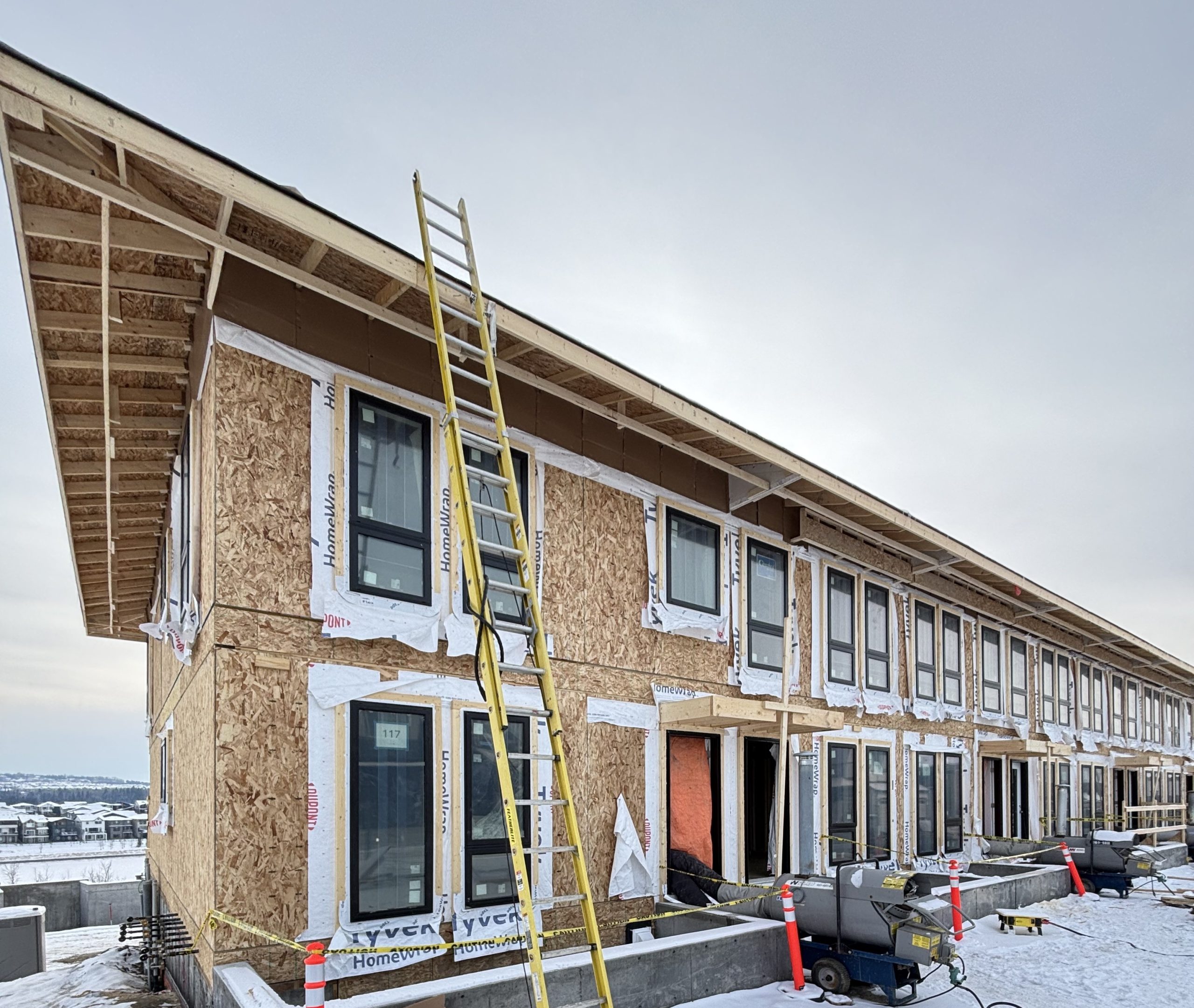 Row of townhomes under construction at ZEN Crimson Ridge in Calgary, showing framed walls, windows, and construction equipment on-site.