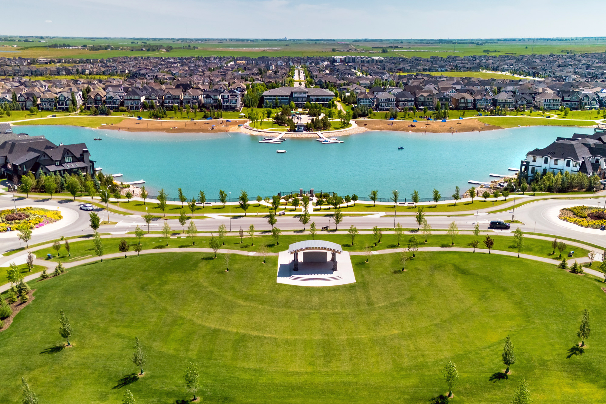 Aerial view of Mahogany Lake in Calgary, featuring a sandy beach, turquoise water, surrounding homes, and a landscaped park with a central pavilion in the foreground.