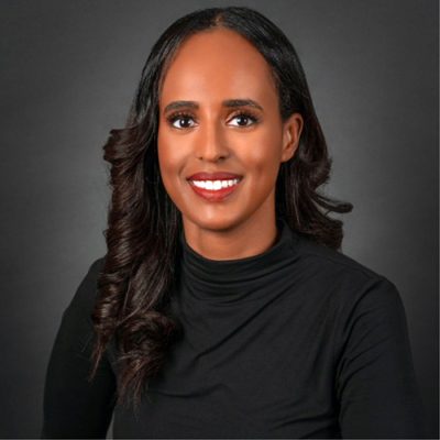 Close-up headshot of a smiling woman with long black hair, wearing a black turtleneck against a grey background.