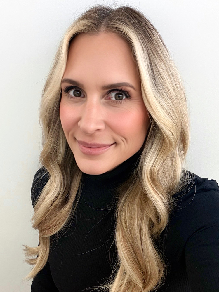 Close-up headshot of a smiling woman with long blonde hair, wearing a black turtleneck against a light background.