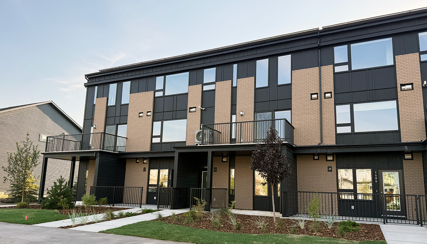 Modern three-story townhouse building with black siding, light brick accents, balconies, and landscaped walkways under a clear blue sky.